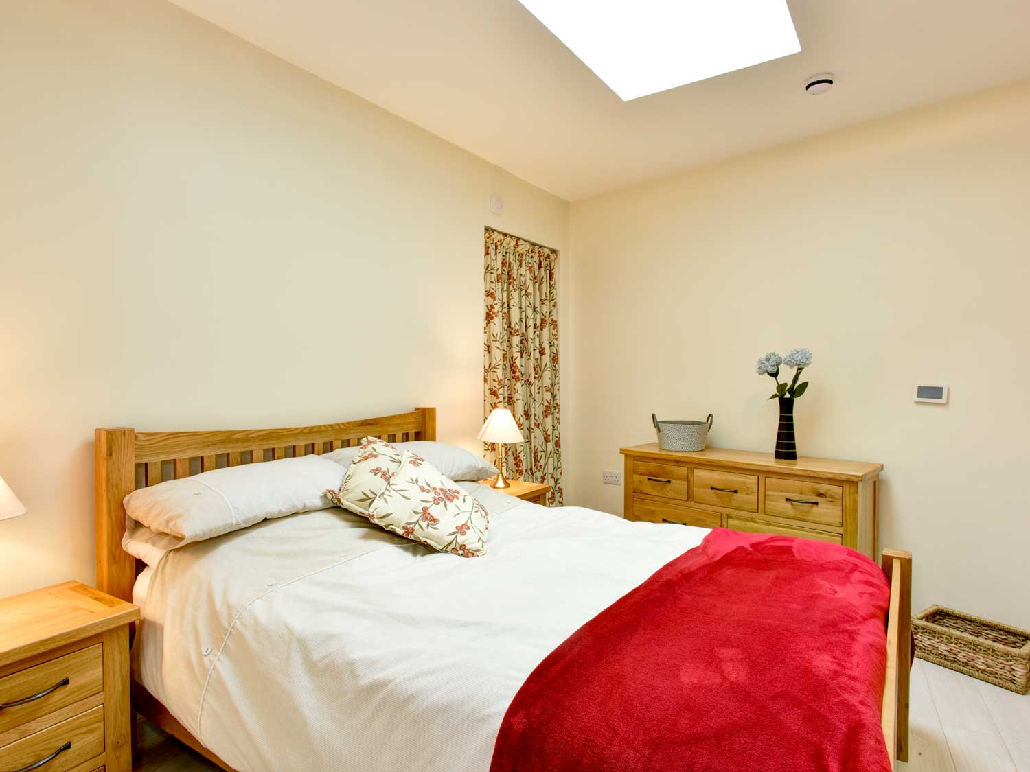 A spacious bedroom with double bed and wooden furnishings. The bed has a patterned pillow and a red blanket thrown over crisp white bed linen